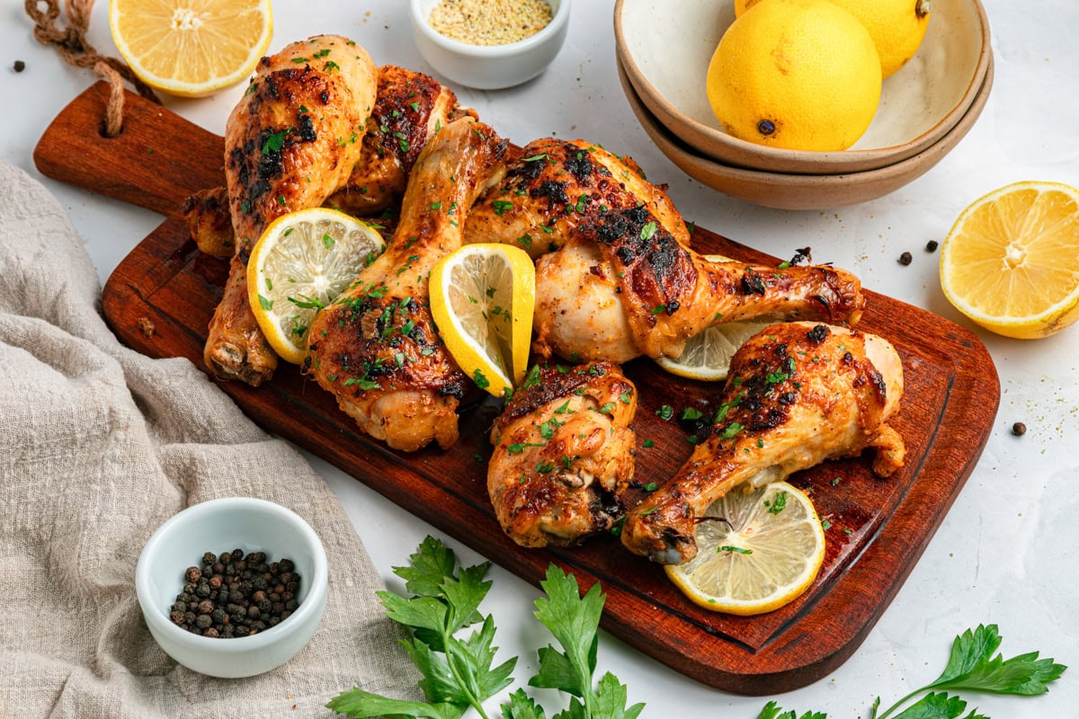 Cooked chicken drumsticks with lemon slices and fresh parsley on a wood cutting board with a handle, with parsley, lemon halved, and seasonings to the side.