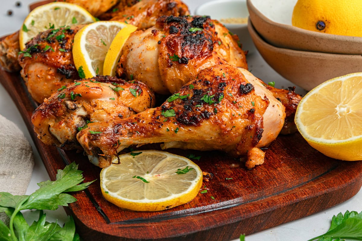 Cooked chicken drumsticks with lemon slices and fresh parsley on a wood cutting board with a handle, with parsley and lemon halves to the side.