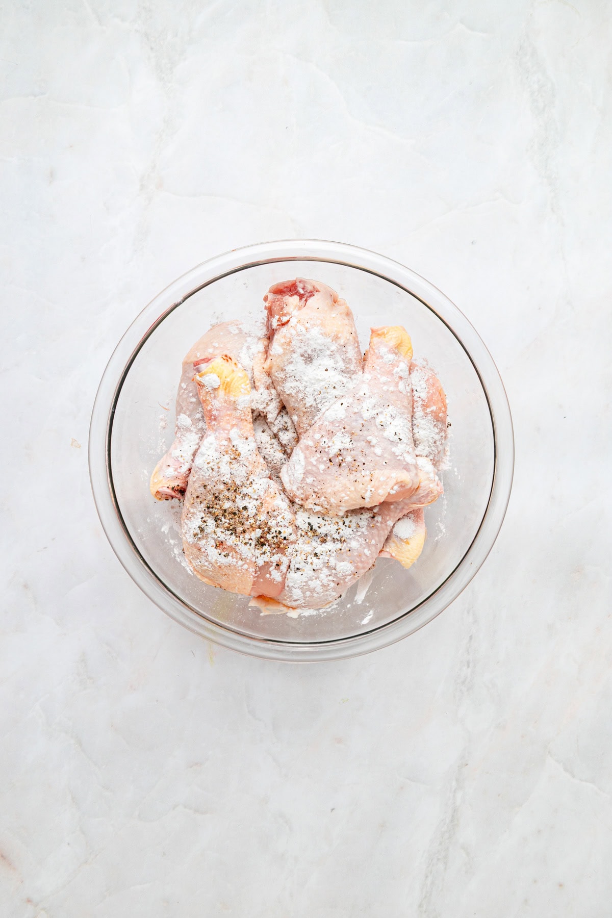 Raw chicken with flour and seasoning in a glass bowl on a white counter.