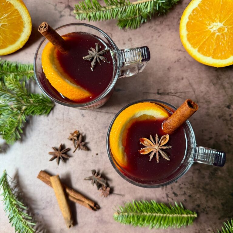 Glass mugs of red liquid with cinnamon stick, star anise, and orange slice, with more spices on the counter along with Christmas greens.