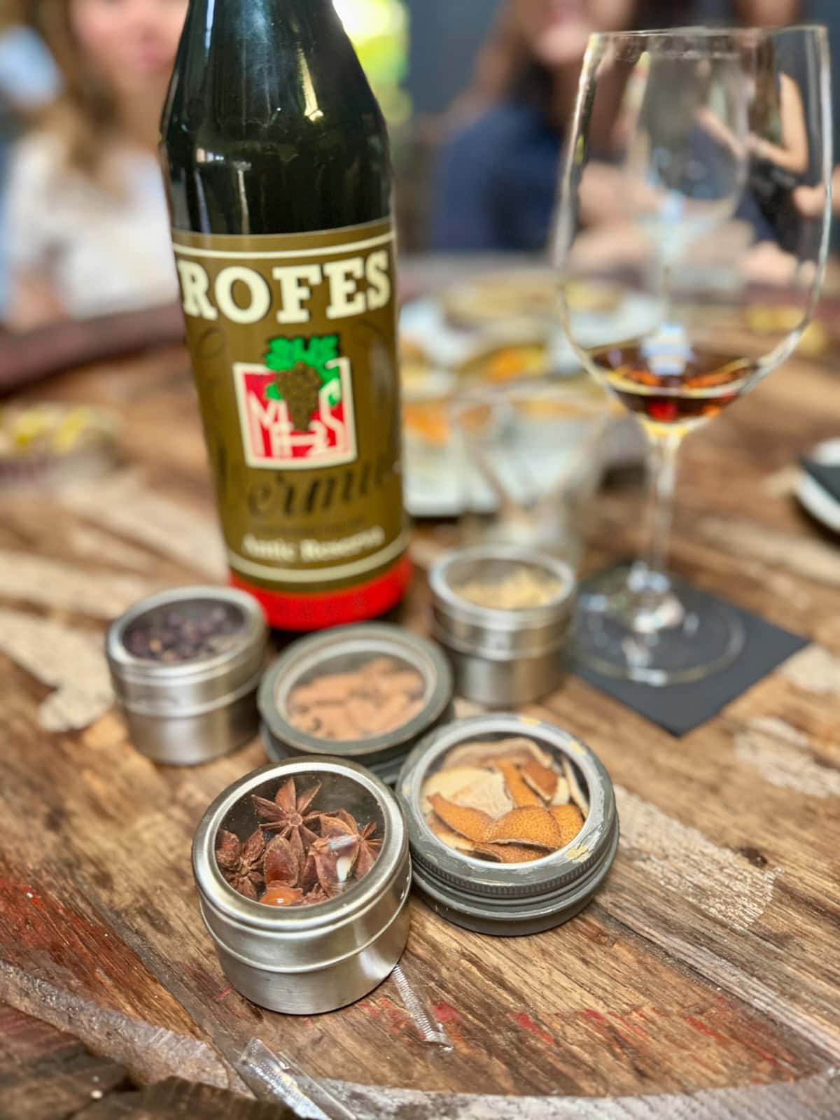 Bottle of wine on a wood barrel table next to a glass with a small amount of wine with small containers of aromatics in front.