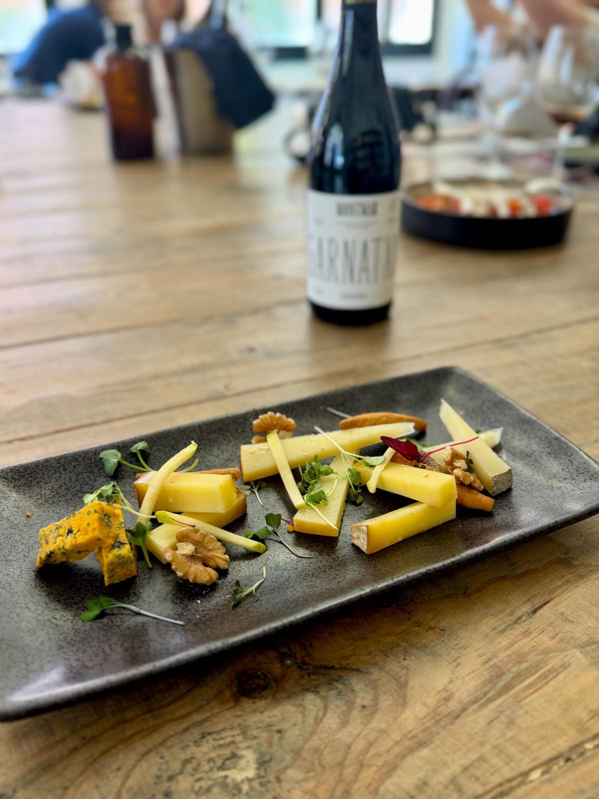 Plate of cheese, nuts, and fresh sprigs of herbs with a bottle of wine in the background.