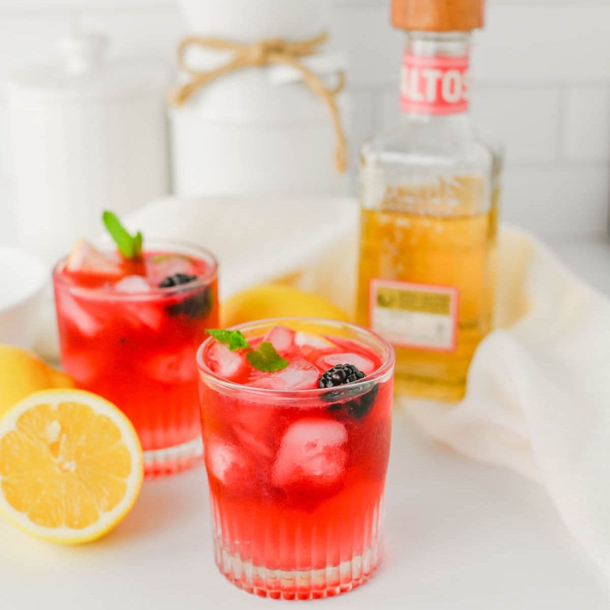 Red colored cocktail with a blackberry and mint sitting in front of a bottle of tequila with a lemon to the side.