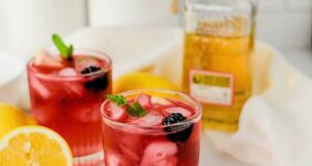 Red colored cocktail with a blackberry and mint sitting in front of a bottle of tequila with a lemon to the side.